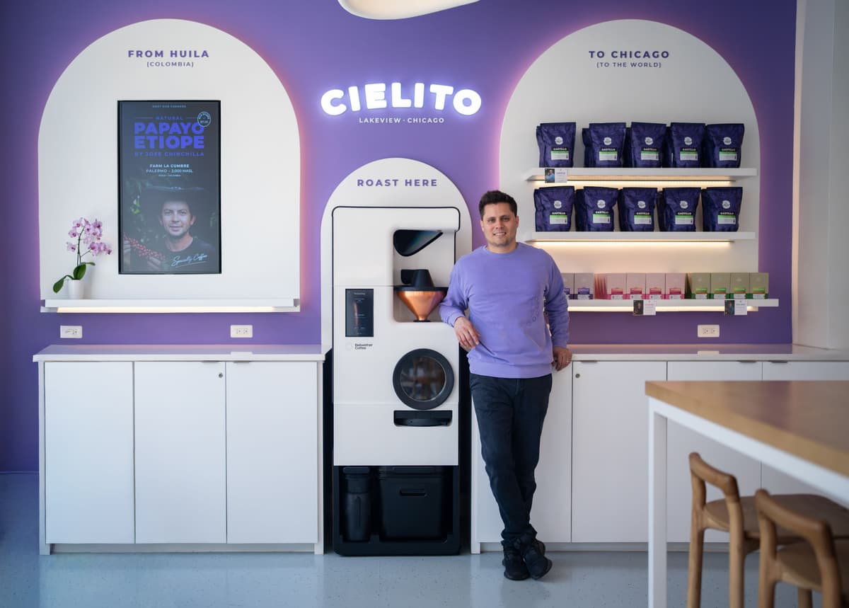 Fernando stands beside the Bellwether Shop Roaster at Cielito in Lakeview, Chicago, under a lit sign reading Roast Here, with shelves of his own Castillo retail bags behind him